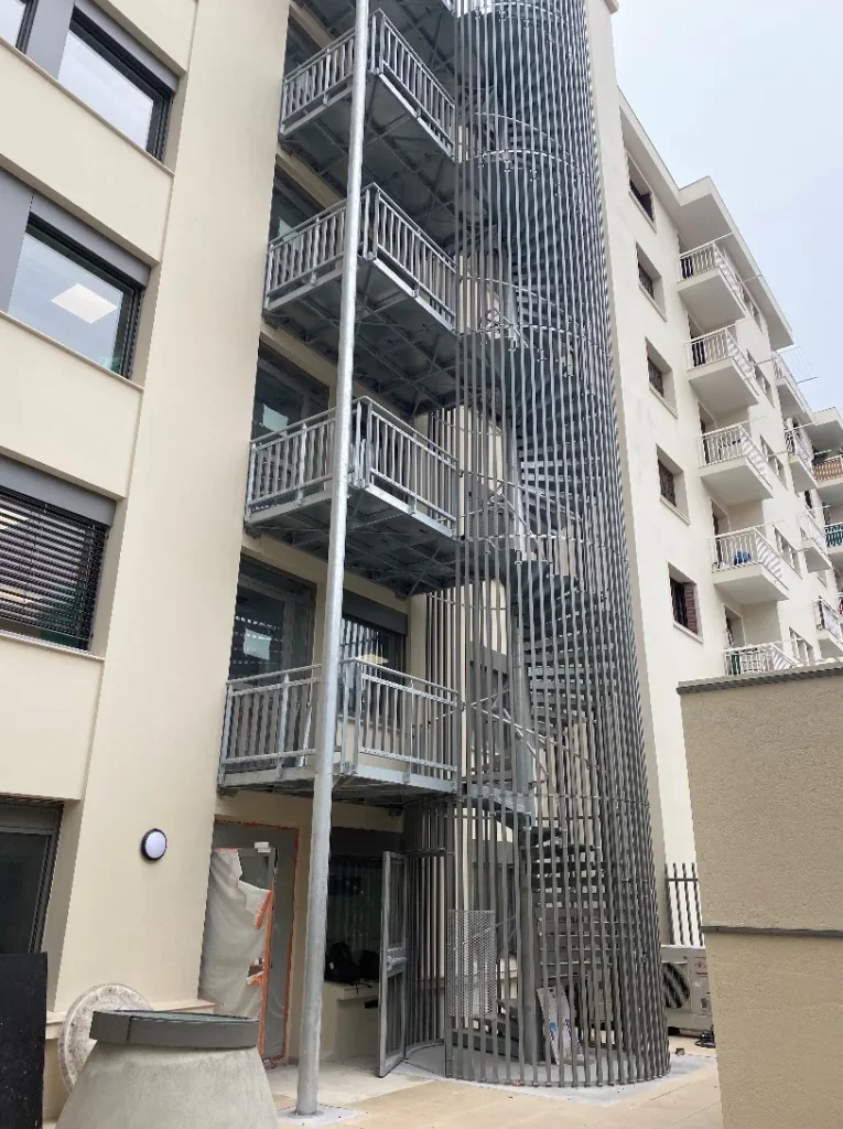 Escalier hélicoïdal métallique galvanisé avec paliers et habillage à barreaudage vertical desservant les étages d'un immeuble